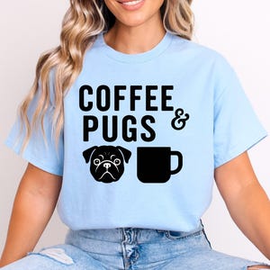Puede incluir: Camiseta azul claro con las palabras "COFFEE PUGS &" en letras negras. Una cara de pug negra y una taza de café negra están impresas debajo del texto. La camiseta es de material suave.