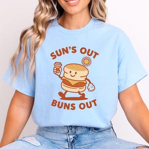 Può includere: T-shirt azzurra con la scritta marrone "SUN'S OUT BUNS OUT". La grafica presenta un biscotto dei cartoni animati sorridente che tiene in mano una tazza di caffè e un sole. La maglietta è di stile casual e confortevole.