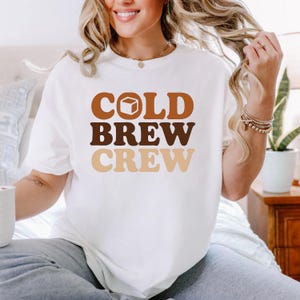 May include: White t-shirt featuring the text "COLD BREW CREW" in brown and beige lettering. The word "COLD" includes a coffee cup graphic. The shirt is a relaxed fit.