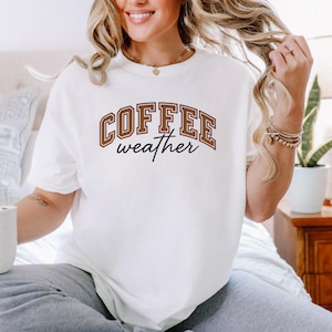 May include: A cream-coloured t-shirt featuring the text "COFFEE weather" in brown block letters. The shirt has a relaxed fit and is made of a soft material. The text is in a collegiate style font.