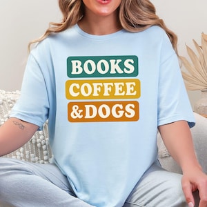 Puede incluir: Camiseta azul claro con la frase "BOOKS COFFEE & DOGS" en un diseño apilado. El texto está en una fuente retro, con cada palabra en un bloque rectangular separado en verde, amarillo y naranja.