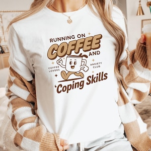 May include: White t-shirt with the text "Running on Coffee and Coping Skills." The design features a cartoon coffee cup character. Additional text includes "Coffee Lovers" and "Anxiety Club."