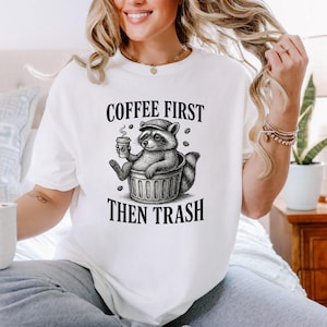 May include: White t-shirt with a black and white illustration of a raccoon in a bin, holding a coffee cup. The text "COFFEE FIRST THEN TRASH" is printed above and below the raccoon design.