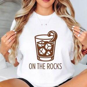 May include: White t-shirt with a brown graphic of a cocktail glass filled with ice cubes and a lemon twist. The words "ON THE ROCKS" are printed below the glass in brown. A casual, everyday wear item.