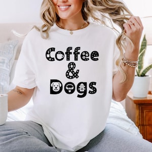Op de afbeelding: Wit T-shirt met de zwarte tekst "Coffee & Dogs". Het woord "Coffee" is versierd met een koffiekopje en bloemmotieven. Het ampersand is gestippeld en "Dogs" bevat een honden gezicht, halsband en brandkraan.
