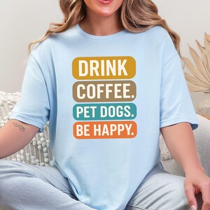 Op de afbeelding: Lichtblauw T-shirt met de tekst "DRINK COFFEE. PET DOGS. BE HAPPY." in een gestapelde vorm. De tekst is in een mix van kleuren, waaronder goud, bruin, teal en oranje. Het shirt heeft korte mouwen en een relaxte pasvorm.