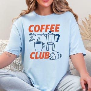 Può includere: T-shirt azzurra con le parole "COFFEE CLUB" in arancione e illustrazioni a tema caffè. Il design include una tazza di caffè, chicchi, una caffettiera e un croissant. È presente anche il testo "all day every day".