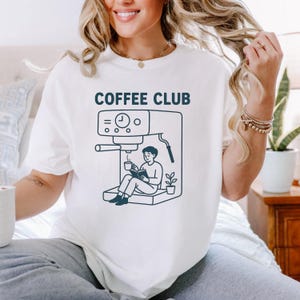Op de afbeelding: Wit T-shirt met de tekst "COFFEE CLUB" boven een donkerblauwe illustratie van een koffiemachine en een persoon die een boek leest terwijl hij een koffiekopje vasthoudt. Het ontwerp is eenvoudig en modern.