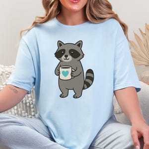 Può includere: T-shirt azzurra con un procione dei cartoni animati che tiene in mano una tazza bianca con un cuore blu. Il procione è grigio con una maschera nera e una coda a strisce. La maglietta è di colore azzurro chiaro.