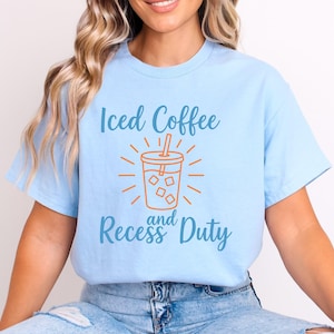 Op de afbeelding: Lichtblauw T-shirt met de tekst "Iced Coffee and Recess Duty" in blauw en oranje. De afbeelding toont een ijscoffiebeker met ijsblokjes en een rietje. Het is een casual kledingstuk.