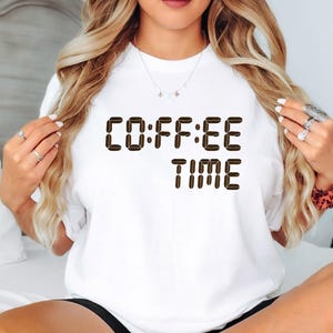 May include: White t-shirt featuring the text "COFFEE TIME" in brown, coffee bean-shaped letters. A simple, casual top for everyday wear.