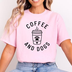 May include: A pale pink t-shirt featuring the text "COFFEE AND DOGS" in black, framing a black graphic of a coffee cup with a paw print and a heart. The shirt is made of a soft material.