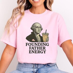 Puede incluir: Camiseta rosa claro con un gráfico de George Washington sosteniendo una taza de café. El texto "FOUNDING FATHER ENERGY" está impreso debajo en negrita negra. La camiseta es de cuello redondo.