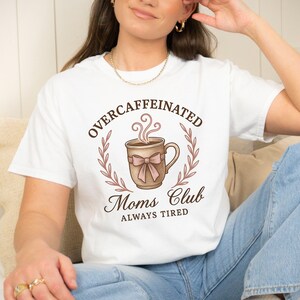 Puede incluir: Camiseta blanca con las palabras "OVERCAFFEINATED Moms Club ALWAYS TIRED" en escritura marrón. El diseño presenta una taza de café con un lazo rosa y vapor saliendo, rodeada por una corona.