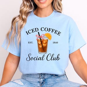 Op de afbeelding: Lichtblauw T-shirt met de tekst "ICED COFFEE Social Club" in zwart script. De afbeelding toont een glas ijskoffie met een rietje en een schijfje sinaasappel, en de tekst "EST. 1840".