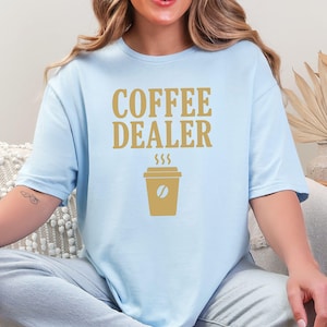 May include: Light blue t-shirt with the words "COFFEE DEALER" in gold lettering. Below the text is a gold coffee cup graphic with a coffee bean and steam rising from the top. The shirt is a casual style.