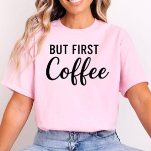 May include: A light pink t-shirt with the text "BUT FIRST Coffee" in black script. The shirt is made of a soft material and has short sleeves. The text is centered on the front of the shirt.