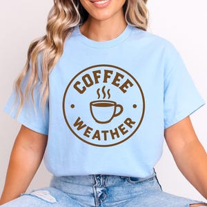 Może przedstawiać: Jasnoniebieska koszulka z brązową okrągłą grafiką. Grafika przedstawia napisy "COFFEE WEATHER" wokół parującego kubka kawy. Codzienny, swobodny element garderoby.