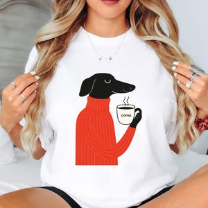 May include: White t-shirt with a cartoon dachshund wearing a red turtleneck sweater, holding a coffee mug. The mug has the word "COFFEE" printed on it. The dog is black with a long snout.