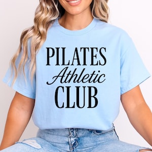 Puede incluir: Camiseta azul claro con el texto negro "PILATES Athletic CLUB". La parte superior es de estilo cuello redondo y parece estar hecha de un material suave. La camisa es de color azul claro liso.