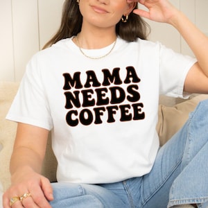 May include: White t-shirt with the text "MAMA NEEDS COFFEE" in a black and brown retro font. The shirt is a casual style, suitable for everyday wear. The design is centered on the front of the shirt.