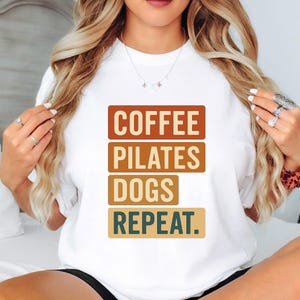 Puede incluir: Camiseta blanca con la frase "COFFEE PILATES DOGS REPEAT" en un diseño apilado. Las palabras están en bloques rectangulares marrones, beige y verdes. Una camiseta sencilla e informal para el uso diario.