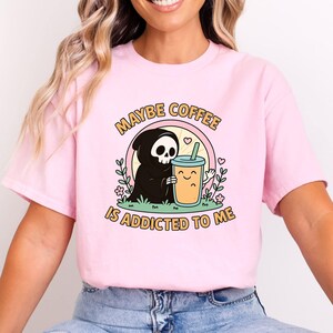 May include: A pale pink t-shirt with a cartoon design of a grim reaper and a coffee cup. The text says "MAYBE COFFEE IS ADDICTED TO ME". The design includes hearts and flowers, with a playful font.
