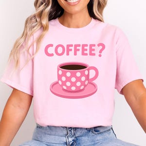 Könnte beinhalten: Hellrosa T-Shirt mit der Aufschrift "COFFEE?" über einer rosa Kaffeetasse mit weißen Punkten auf einer passenden Untertasse. Die Tasse ist mit dunkelbrauner Flüssigkeit gefüllt.