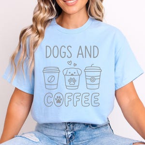 以下が含まれることがあります： 「DOGS AND COFFEE」の文字がグレーでプリントされた水色のTシャツ。コーヒー豆、犬の足跡、円のイラストが描かれた3つのコーヒーカップのデザイン。犬とコーヒー好きへのプレゼントに最適です。