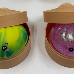 Puede incluir: Dos juguetes novedosos, cada uno en un recipiente marrón con tapa. Uno es un remolino verde y amarillo con una cara de dibujos animados. El otro es una bola de purpurina rosa con una cara de dibujos animados.