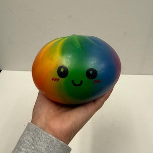 Puede incluir: Un juguete de peluche redondo, de color arcoíris, con una cara sonriente. El juguete tiene ojos negros, mejillas rosas y una boca curvada. Los colores van del rojo al naranja, amarillo, verde y azul. El juguete se sostiene en una mano.