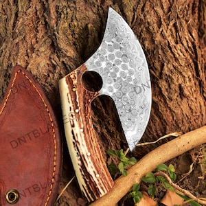 May include: A small, hand-forged axe with a brown and white antler handle. The axe head has a hammered finish and a circular hole in the center. The axe is resting on a piece of wood and a brown leather sheath.