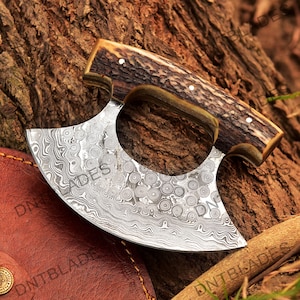 May include: A curved, stainless steel knife with a damascus pattern and a brown antler handle. The knife is designed for chopping and has a rounded blade.