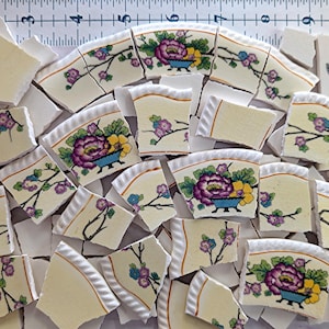 May include: A collection of broken ceramic pieces with floral designs. The fragments feature a cream-colored background with purple, blue, and yellow flowers. Some pieces have a blue bowl with a large purple flower. The pieces are scattered.