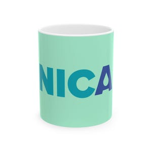 Puede incluir: Una taza de cerámica verde menta con el texto NICA impreso en letras mayúsculas negritas sin remates. Las letras N, I y C son de color azul cerceta, mientras que la letra A es de color azul marino oscuro con una pequeña gota blanca dentro.