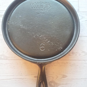 May include: A black cast iron skillet with a long handle. The skillet has the text "WAGNER WARE SIDNEY -0-" and the number "6" embossed on the surface. The skillet is round with a raised edge.