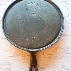 May include: A black cast iron griddle pan with a long handle. The pan has a circular cooking surface and features the stamped text "WAGNER SIDNEY" and the number "7". The pan is designed for stovetop cooking.