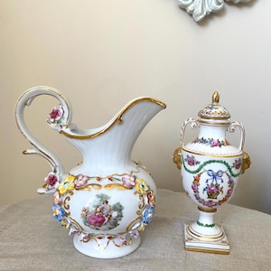Pode incluir: Jarro e urna com tampa de porcelana branca com detalhes florais e dourados. O jarro apresenta uma alça com um adorno floral e uma cena pintada. A urna tem duas alças e uma base quadrada, com um design floral e de laço.
