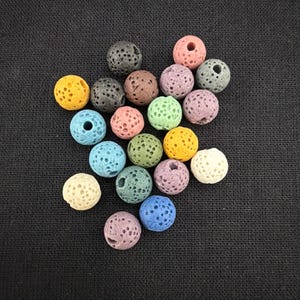 May include: A collection of round, porous lava beads in a variety of colors including yellow, blue, green, pink, purple, brown, gray, black, and off-white. Each bead features a textured surface with small holes and a central hole for threading.