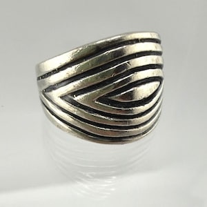 May include: A silver-toned metal ring featuring a series of raised, horizontal, and curved ridges that wrap around the band. The design consists of multiple parallel lines that create a layered, textured appearance across the surface of the ring.