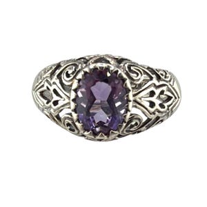 May include: A silver ring featuring an oval-cut purple gemstone set in a prong setting. The band is crafted with intricate openwork metal patterns that extend from the central stone, showcasing a detailed filigree design throughout the metalwork.