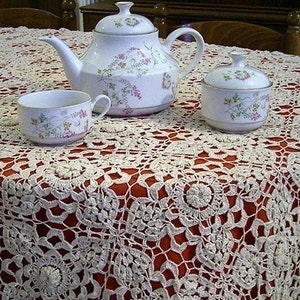 May include: A white teapot with floral designs sits on a white crocheted tablecloth with a red background. A white sugar bowl with a lid and a white teacup with floral designs are also on the tablecloth.