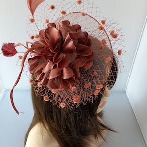 May include: A rust-coloured fascinator with a floral design and a veil. The headpiece features a large, layered flower, feathers, and a net veil with small, round embellishments. The accessory is designed to be worn on the head.