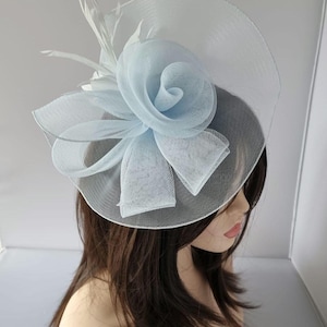 Op de afbeelding: Een lichtblauwe en witte fascinator met een bloemenontwerp. Het hoofddeksel heeft een grote, gelaagde bloem en een doorschijnende sluier. Witte veren accentueren het ontwerp en creëren een elegante uitstraling.