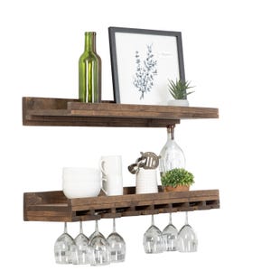 May include: Two-tier, dark-stained wooden wine rack with stemware holders. The top shelf holds a green wine bottle, framed botanical print, and small succulent. The bottom shelf displays wine glasses, white mugs, and decorative items.