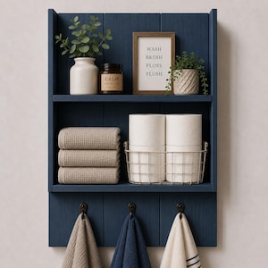 May include: A dark blue wooden wall shelf with two tiers and three hooks. The top shelf holds a white vase with greenery, a brown candle labeled CALM, a framed sign reading WASH BRUSH FLOSS FLUSH, and a potted plant. The lower shelf holds three folded beige towels and a wire basket with two toilet paper rolls. Three towels hang from the hooks.