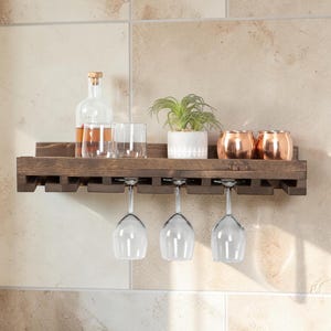 May include: A dark brown wooden wall-mounted shelf featuring a top surface for storage and a lower rack with slots for hanging three clear glass wine glasses. The shelf holds a glass bottle, two tumblers, a small potted plant, and two copper mugs.