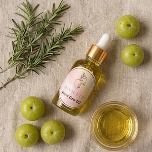 Può includere: Una bottiglia di vetro trasparente di olio per capelli DivieGlo Hair Gro Oil con contagocce e tappo dorato, accanto a uva spina verde, una piccola ciotola di vetro con olio e rametti di rosmarino, su una superficie di lino beige.