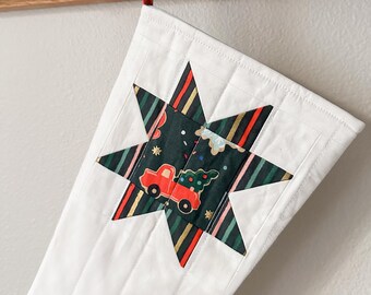 Op de afbeelding: Witte, gewatteerde kerstsok met een stervormige patch met een rode vrachtwagen die een kerstboom vervoert. De stervormige patch is gemaakt van groene, zwarte, gouden, rode en roze stof.