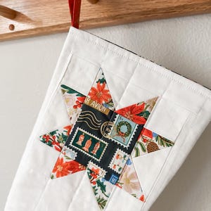 May include: White quilted Christmas stocking with a star-shaped patch made of colorful fabric featuring a vintage postage stamp design. The stocking is hanging on a wooden hook.